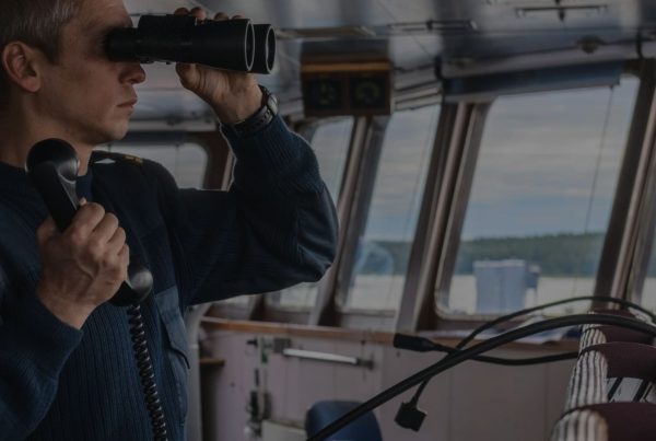 Ship officer looking through binoculars while holding a corded phone on the bridge of a vessel, with large windows overlooking the sea and distant land.